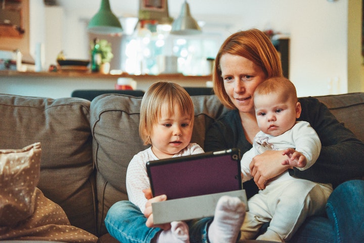 A mother sits on a couch with two young children, pointing at a tablet screen