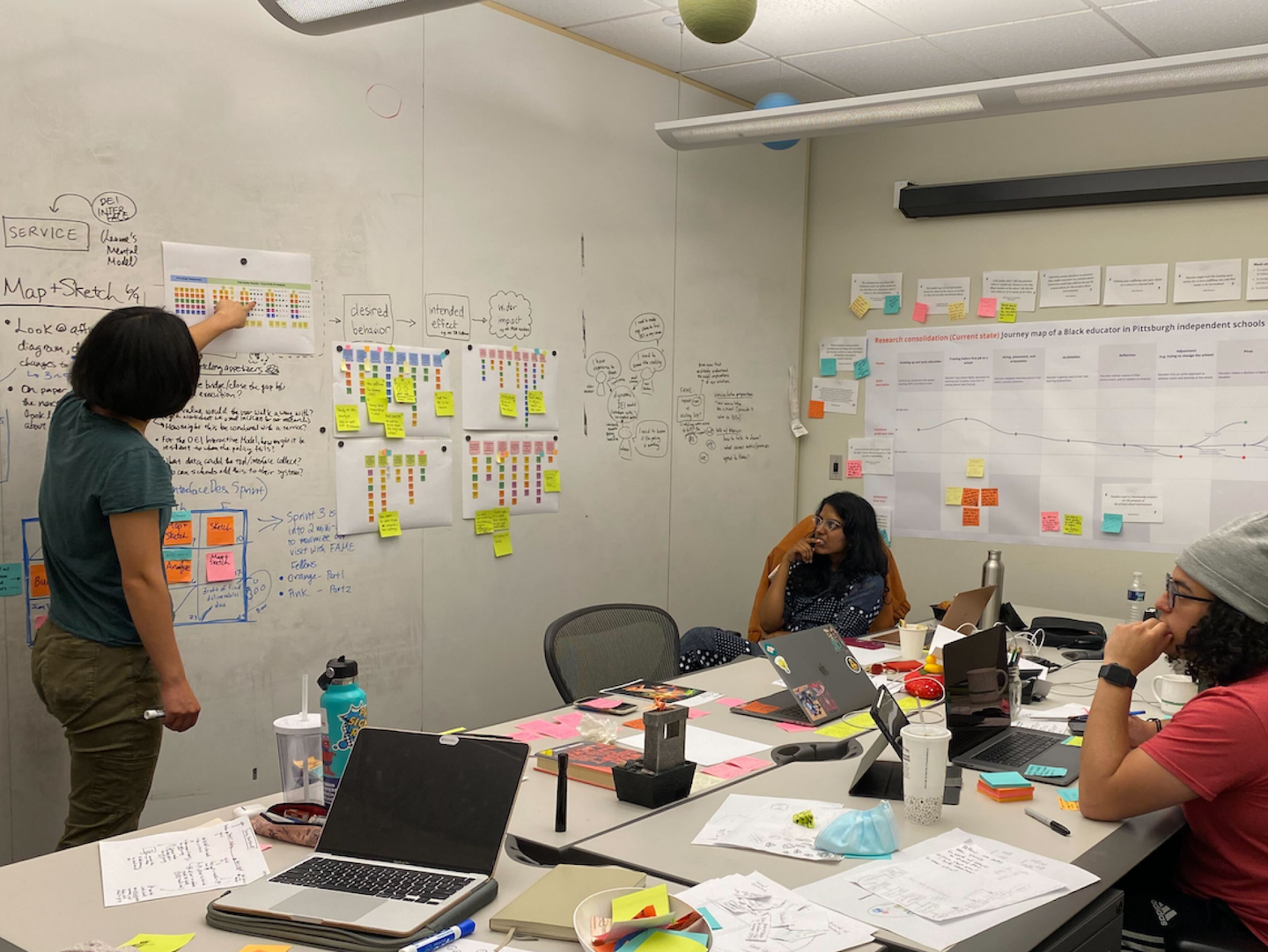 Martina points at a printout on a board filled with design notes, while her team listens attentively