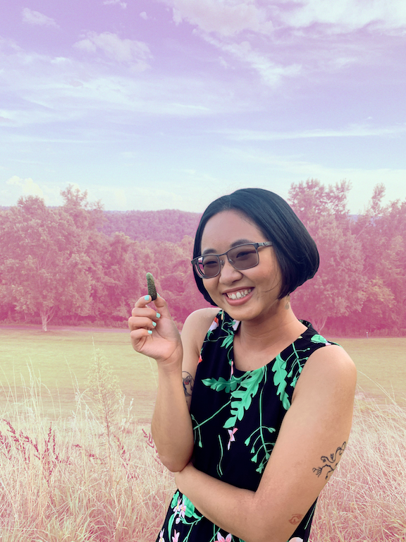 A photo of Martina in a black dress with bright green floral patterns, holding a small pickle and smiling